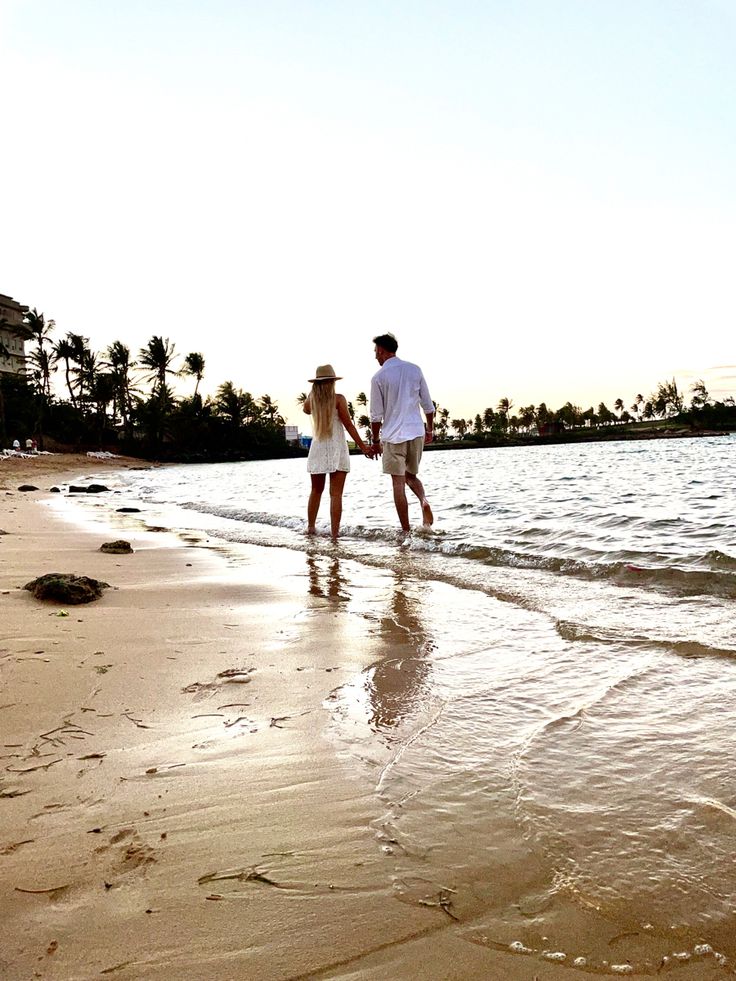 a man and woman holding hands walking along the beach with footprints in the sand as the sun sets