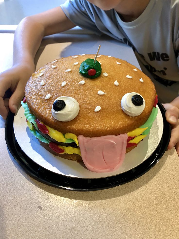 a young boy sitting at a table in front of a large hamburger cake with googly eyes