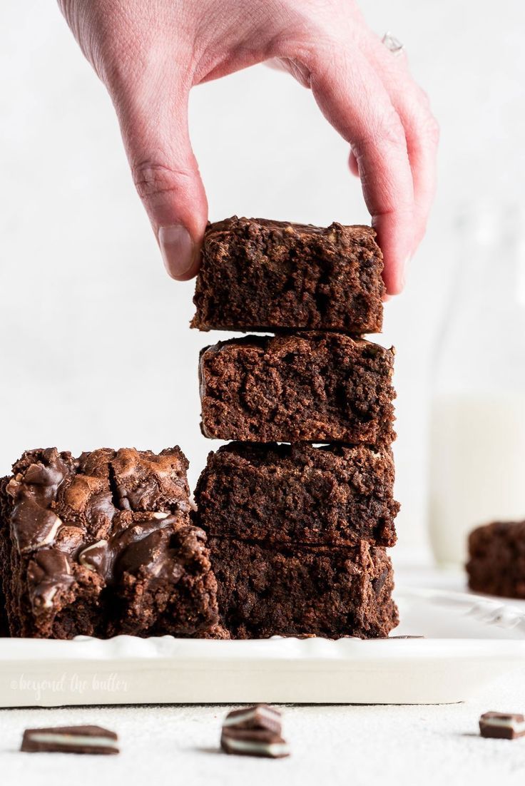 a hand picking up some chocolate brownies from a white plate with milk in the background