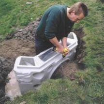 a man standing over a white toilet in the middle of some dirt and green grass