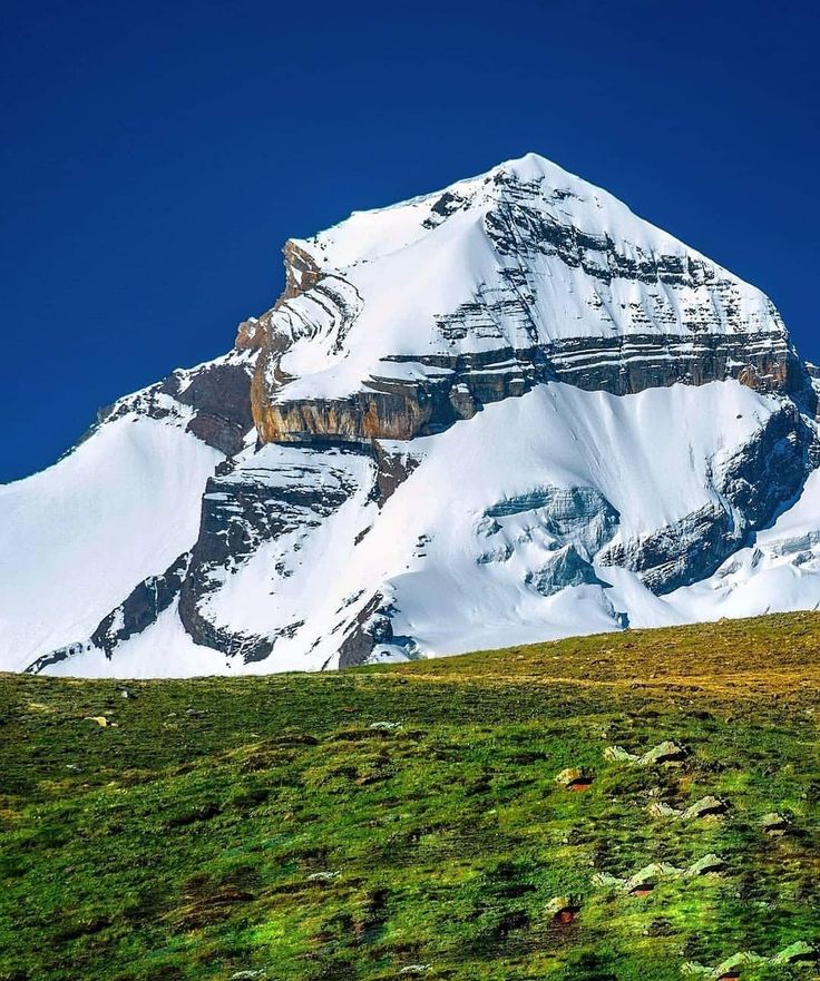 a snow covered mountain with green grass on the ground and blue sky in the background