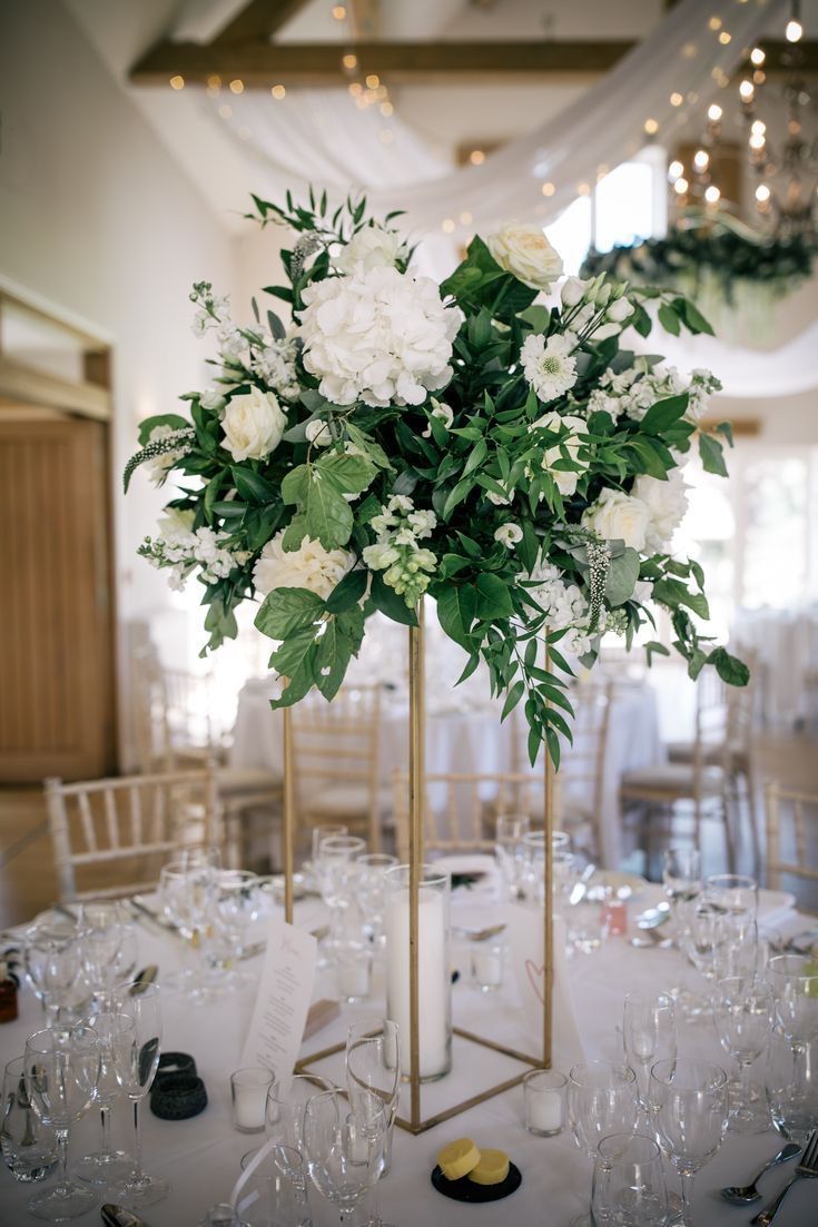 there is a tall vase with white flowers in it on top of a table filled with wine glasses