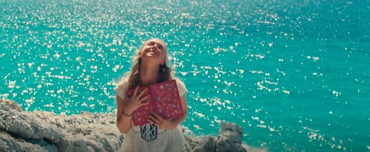 a woman standing on top of a cliff next to the ocean holding a pink bag