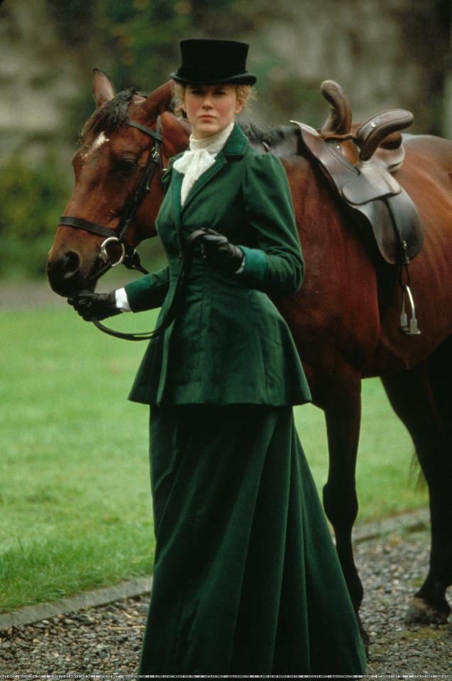 a woman standing next to a brown horse wearing a green dress and top hat with her hand on the bridle