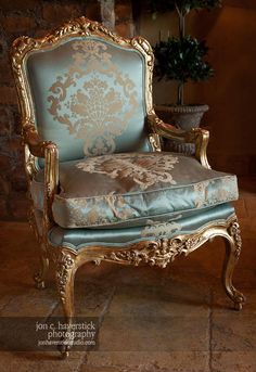 an ornate gold and blue chair sitting in front of a potted plant on a stone floor