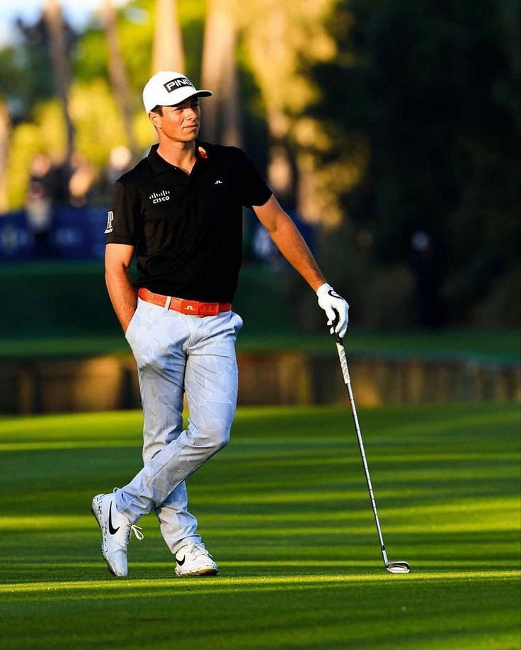 a man in black shirt and white hat holding a golf club while walking on grass