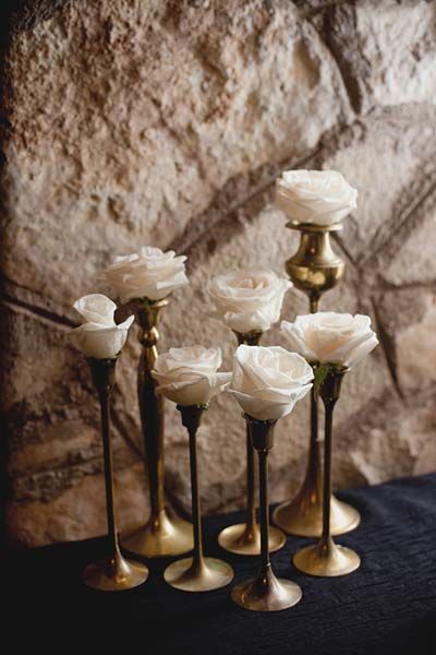 five white roses are in gold vases on a black tablecloth with a stone wall behind them