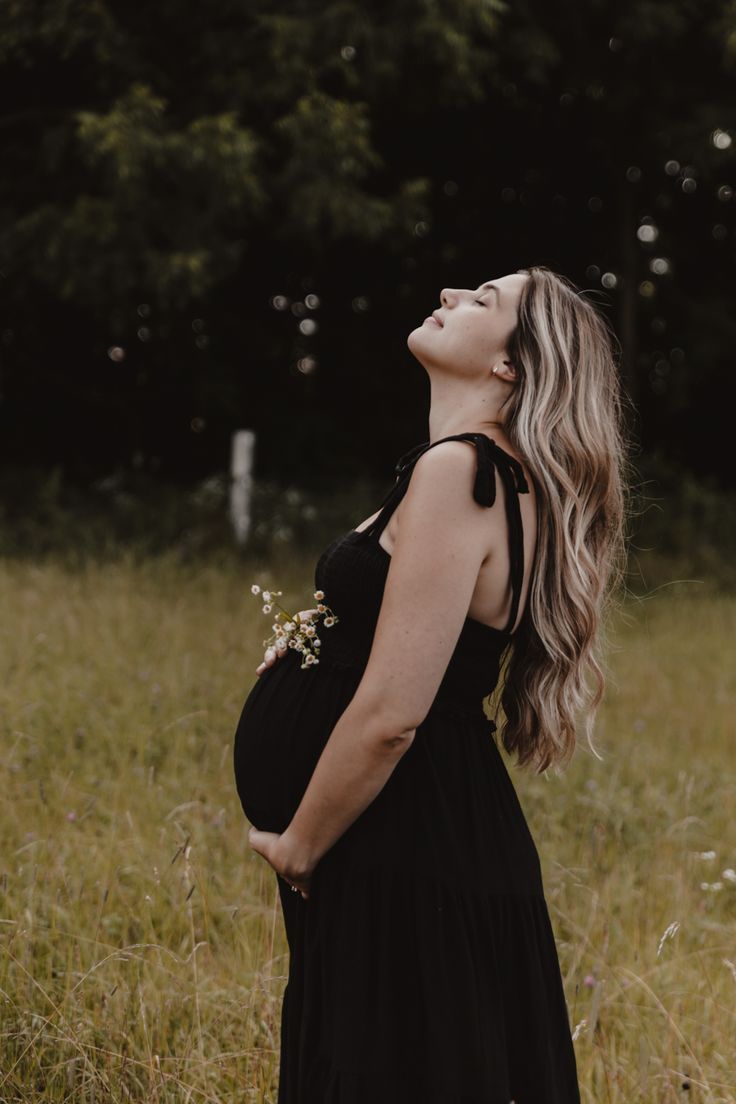 a pregnant woman in a black dress standing in tall grass looking up at the sky