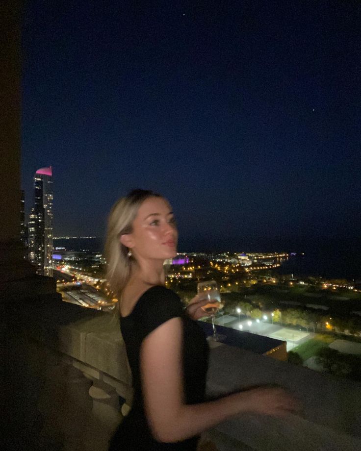 a woman standing on top of a balcony next to a tall building at night with city lights in the background