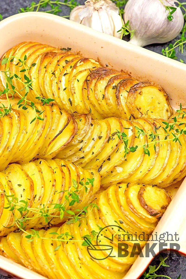 baked potatoes in a casserole dish with herbs on top and garlic sprigs