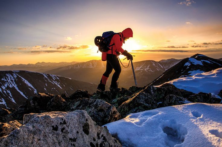 a man hiking up the side of a snow covered mountain at sunset with ski poles in hand