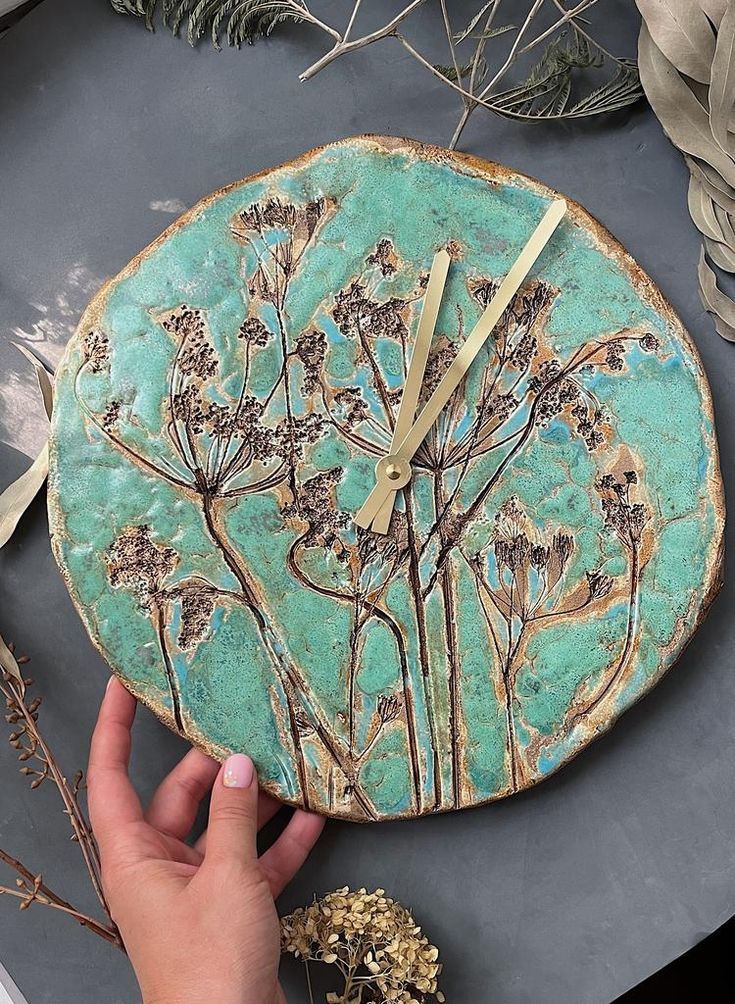 a clock with flowers painted on it in the shape of a tree and leaves, is being held by a person's hand