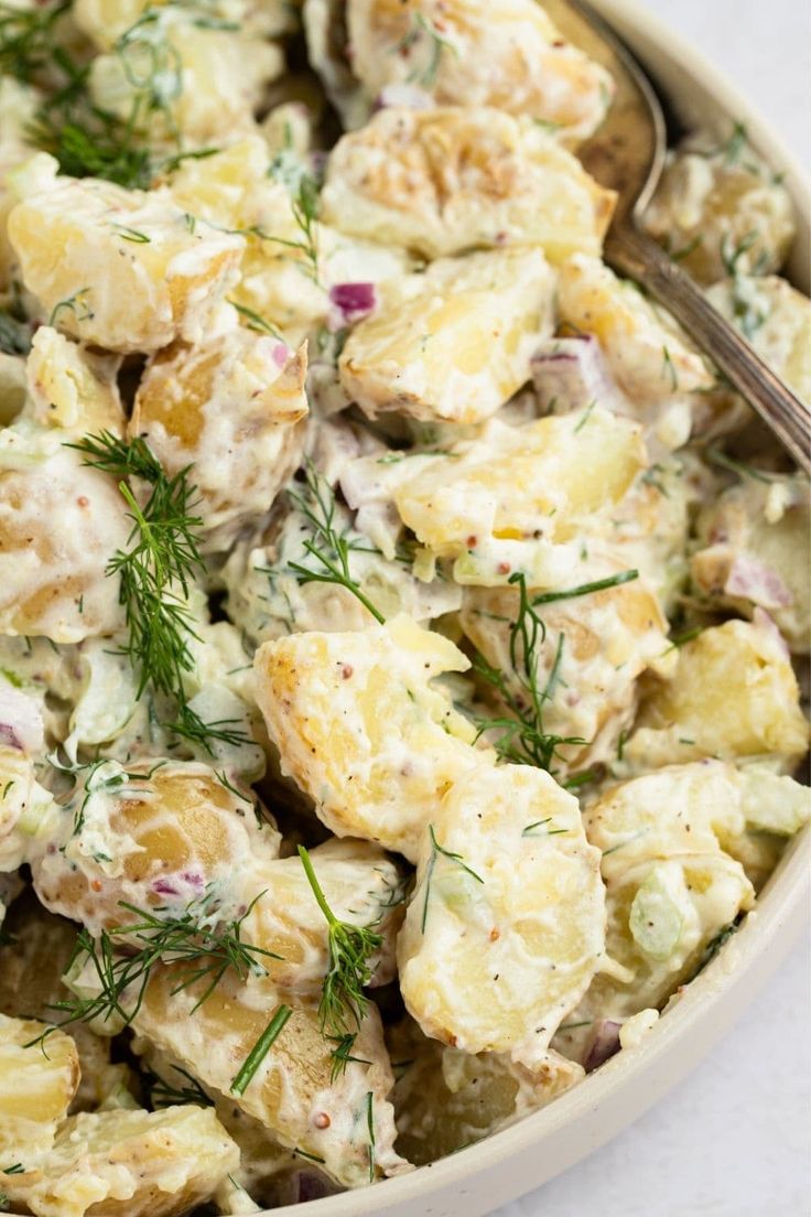 a bowl filled with potato salad on top of a white tablecloth next to a spoon