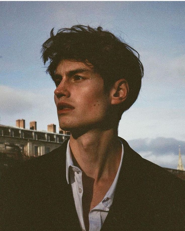 a man in a black suit and white shirt looking off into the distance with buildings in the background