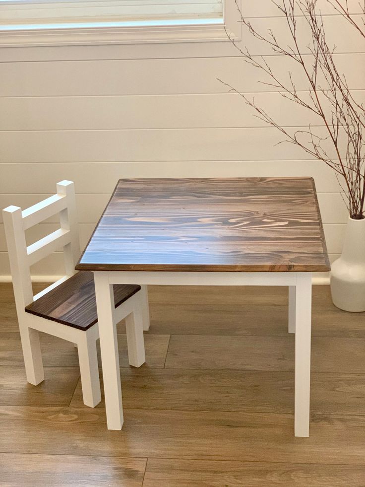 a wooden table and two white chairs in front of a vase with flowers on it