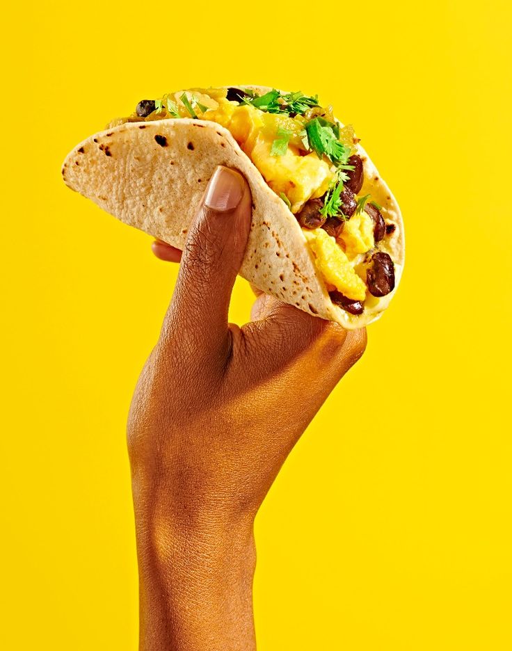 a hand holding a tortilla with eggs and black olives on it against a yellow background