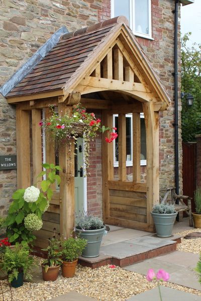 a small wooden building with potted plants and flowers in the foreground, next to a brick wall