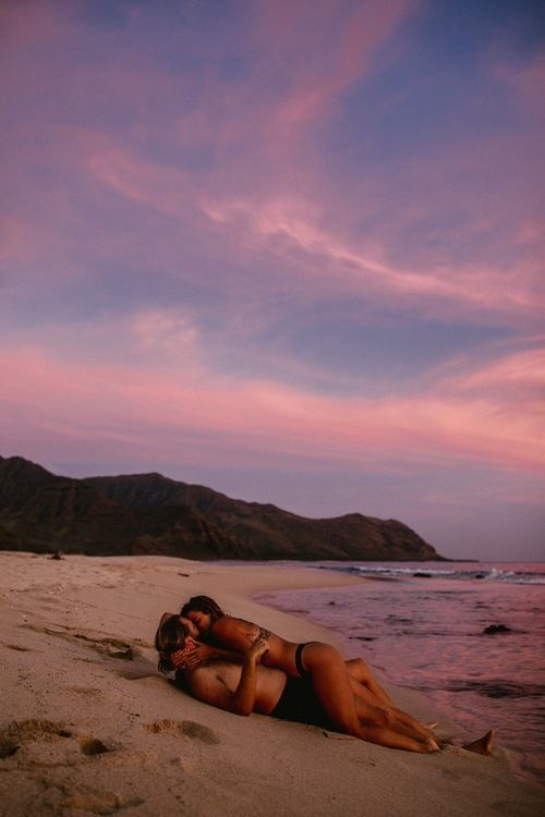 a woman laying on top of a sandy beach under a pink and blue sky next to the ocean