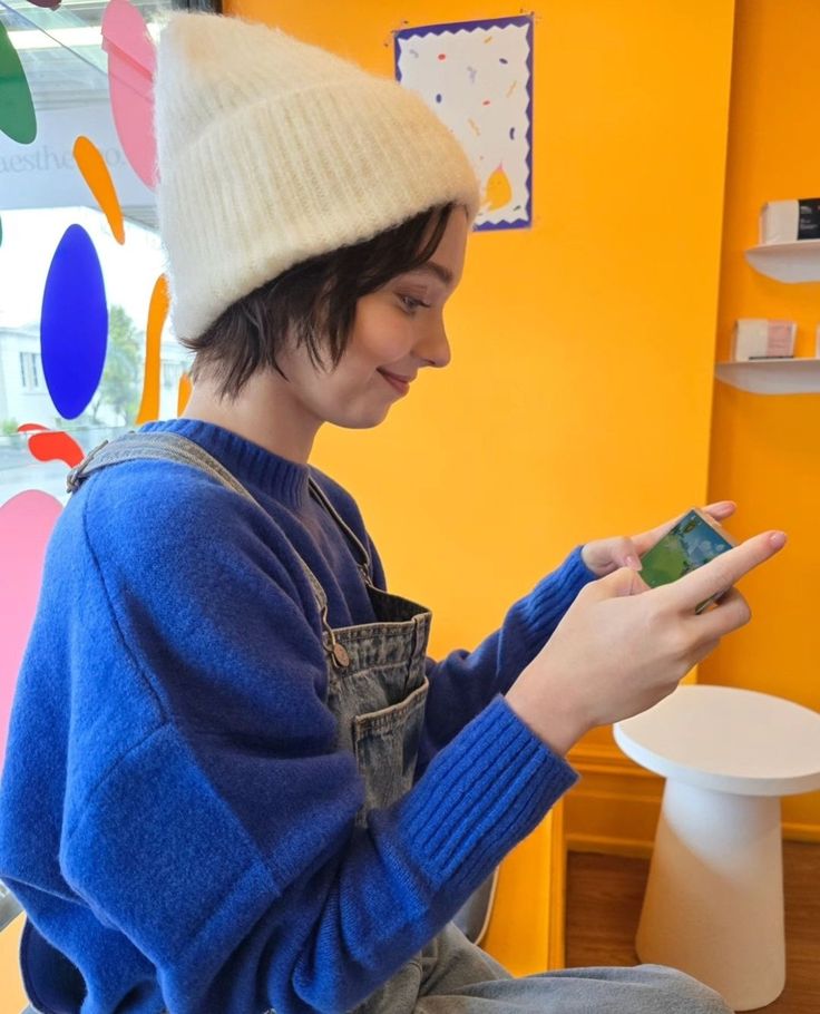 a young boy sitting on the floor looking at his cell phone while wearing a hat