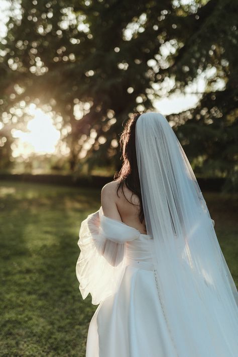 a woman in a white wedding dress and veil standing on the grass with her back to the camera