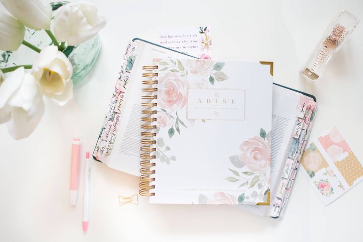 an open planner book sitting on top of a white table next to flowers and pens