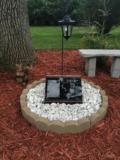 a memorial sits in the middle of a garden with rocks and gravel around it, next to a tree