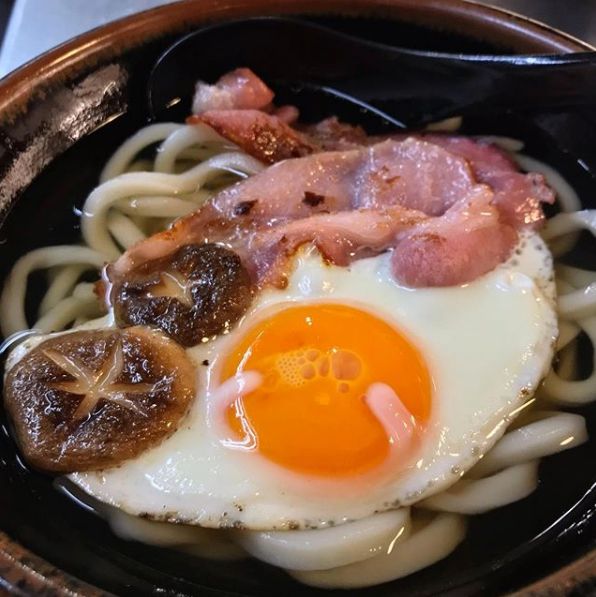 an egg, bacon and noodles dish in a black bowl