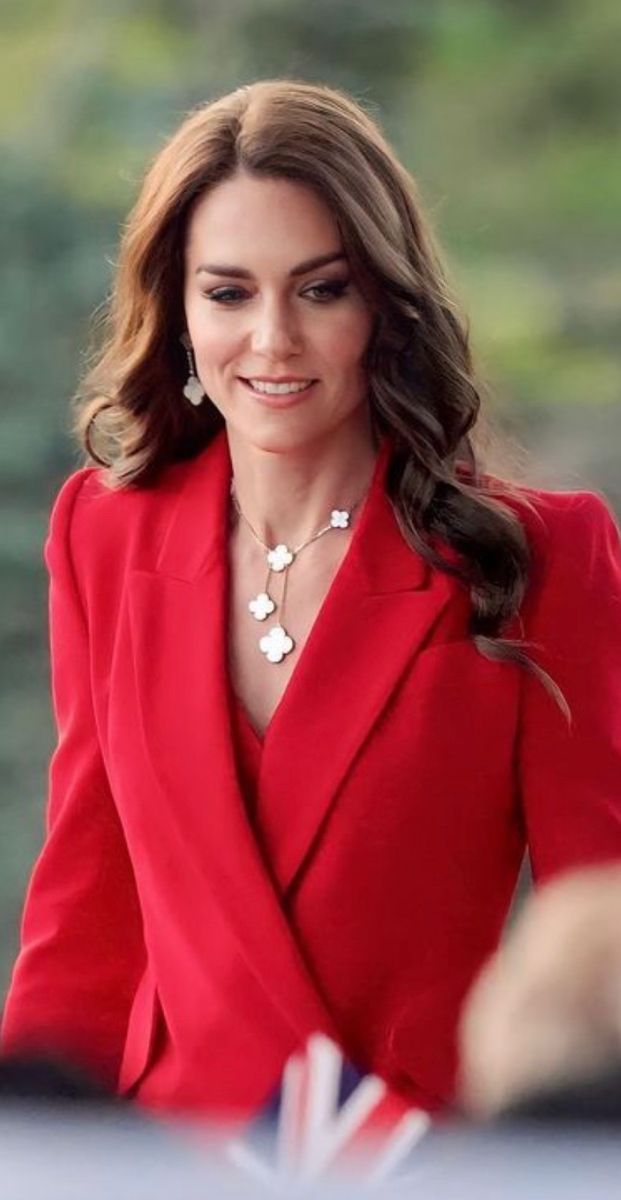 a woman in a red suit and white necklace smiles at the camera as she walks