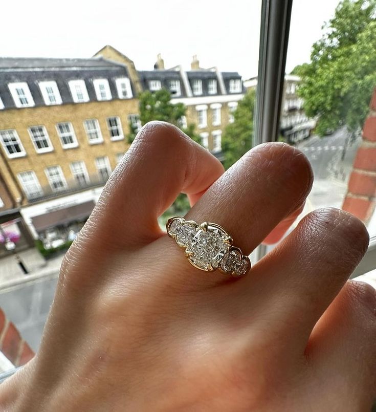 a woman's hand holding a three stone ring in front of a window overlooking the city