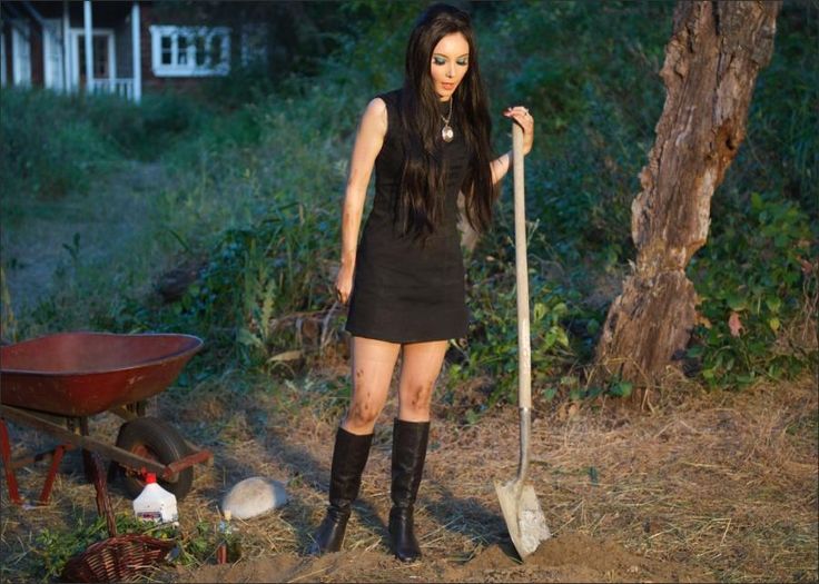 a woman in black dress holding a shovel and standing next to a wheelbarrow