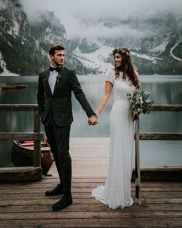 a bride and groom holding hands on a dock in front of a lake with mountains