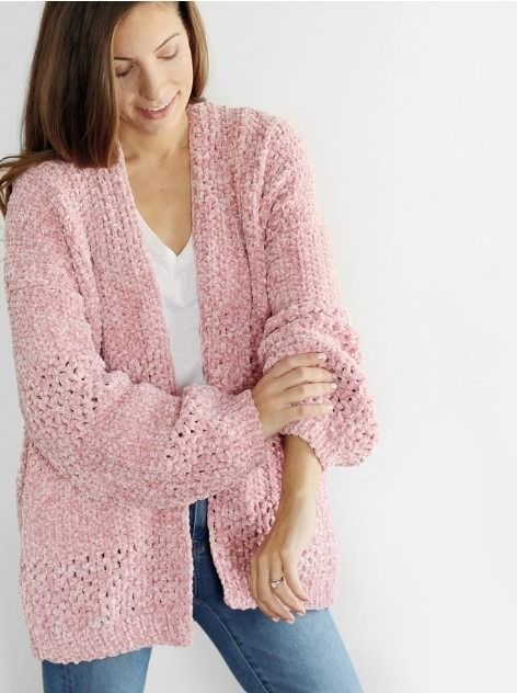 a woman wearing a pink knitted cardigan and jeans standing in front of a white wall