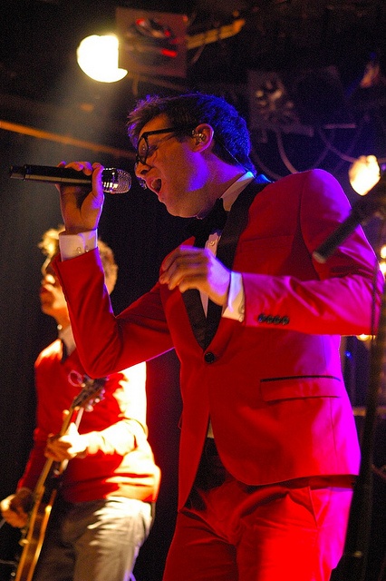 a man in a red suit singing into a microphone while two other men are on stage behind him