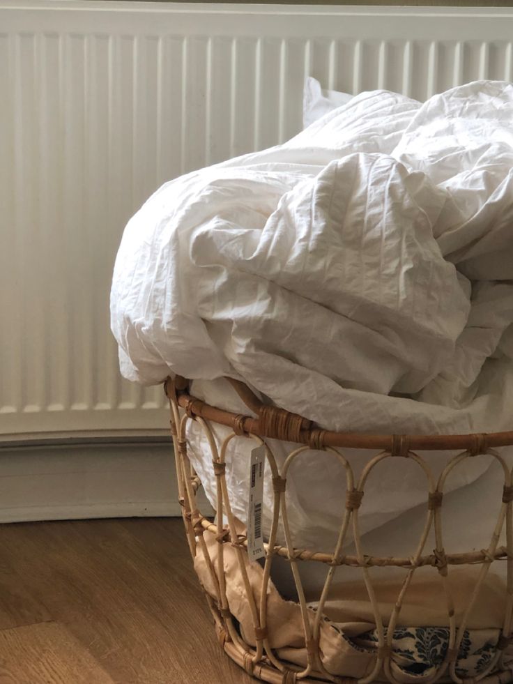 a basket filled with lots of white sheets on top of a hard wood floor next to a radiator