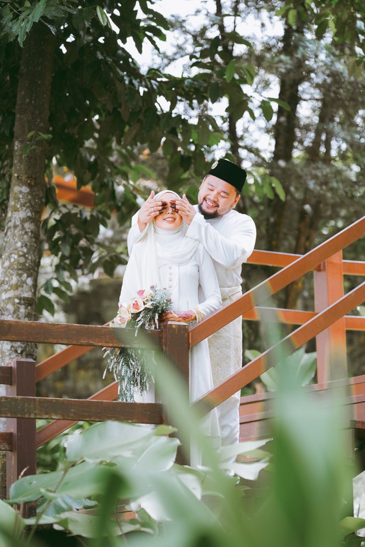 a man and woman standing on a wooden bridge in the woods holding hands up to each other