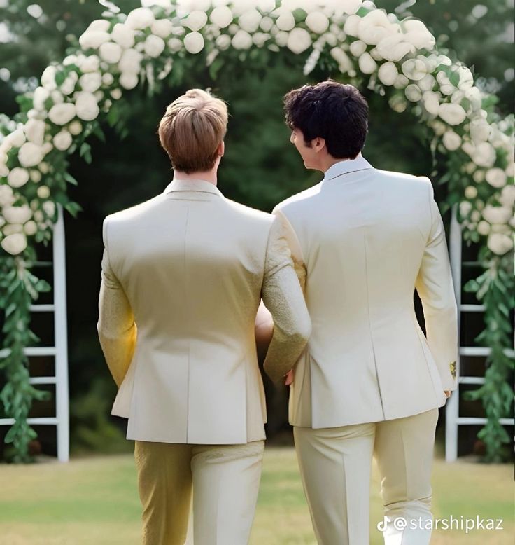 two men in beige suits standing under an arch with white flowers and greenery on it