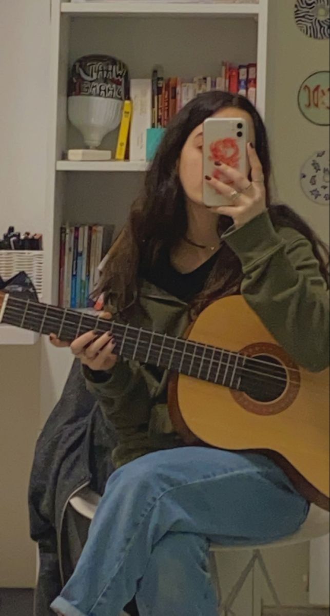 a woman sitting on a chair holding a guitar and taking a selfie with her phone