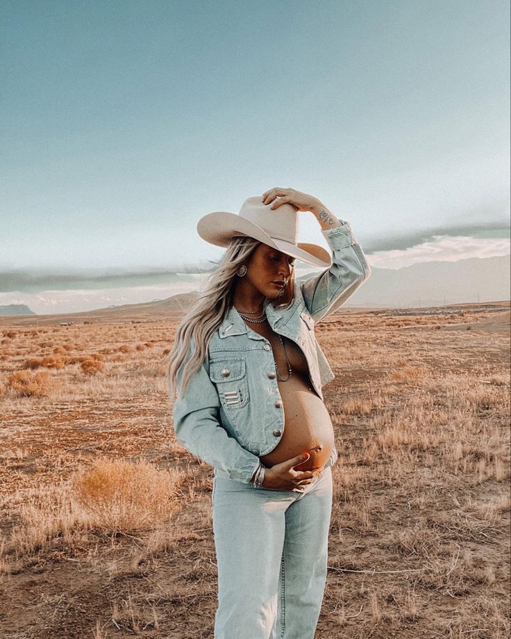 a pregnant woman standing in the middle of a field wearing a cowboy hat and jeans