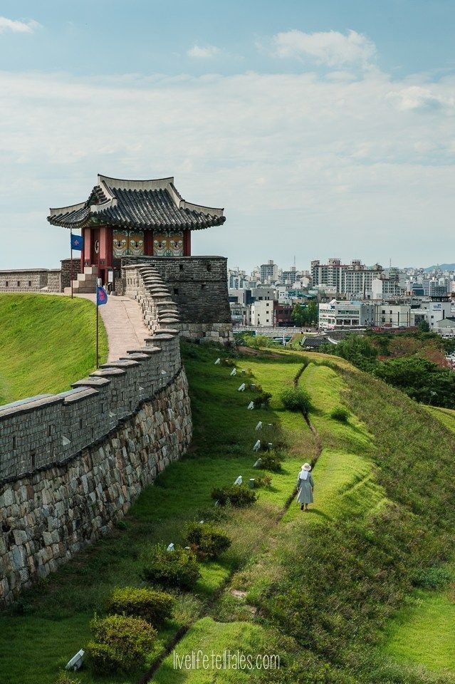 a person standing on top of a grass covered hill next to a wall and building