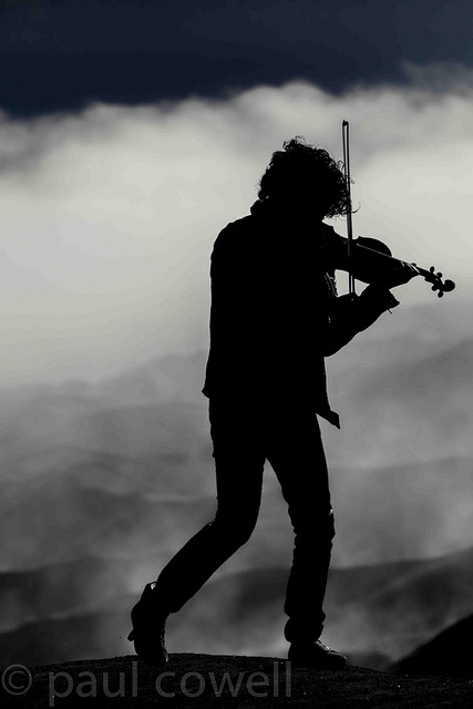 the silhouette of a man playing violin on top of a mountain with clouds in the background