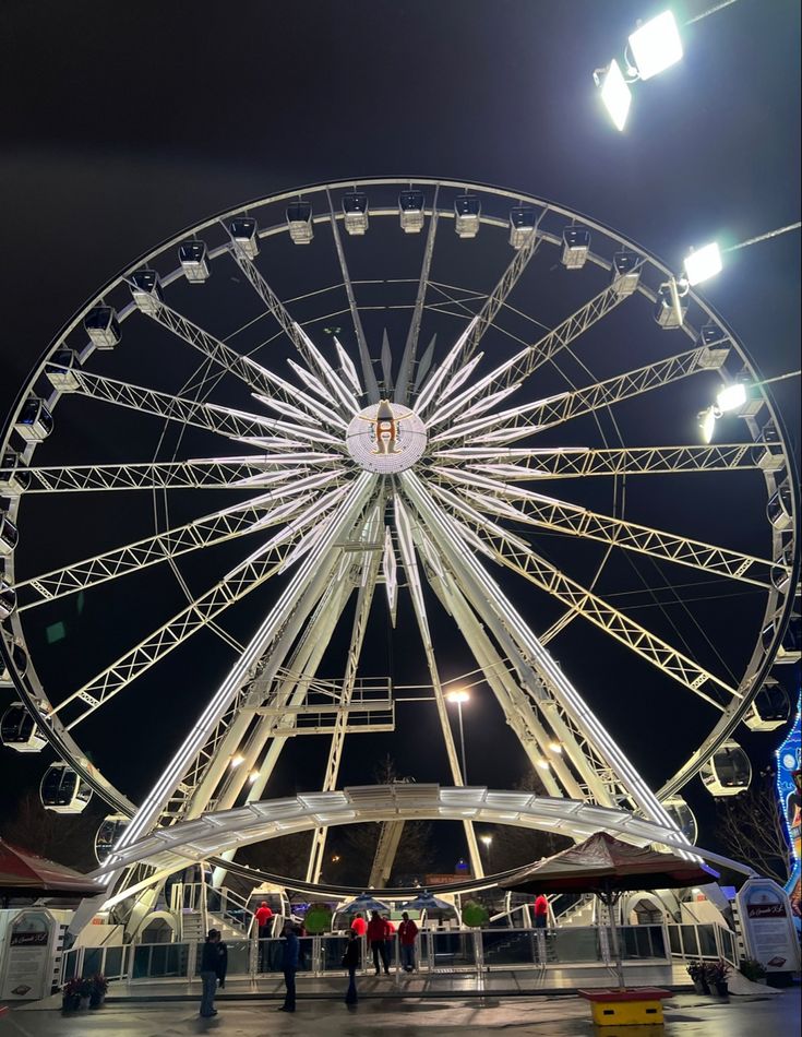 the ferris wheel is lit up at night time with people standing around and looking on