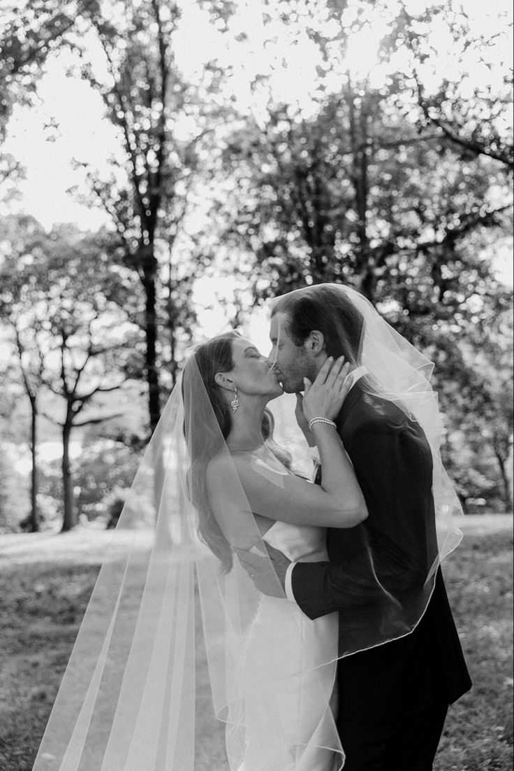 a bride and groom kissing in front of the trees at their wedding day, black and white photograph