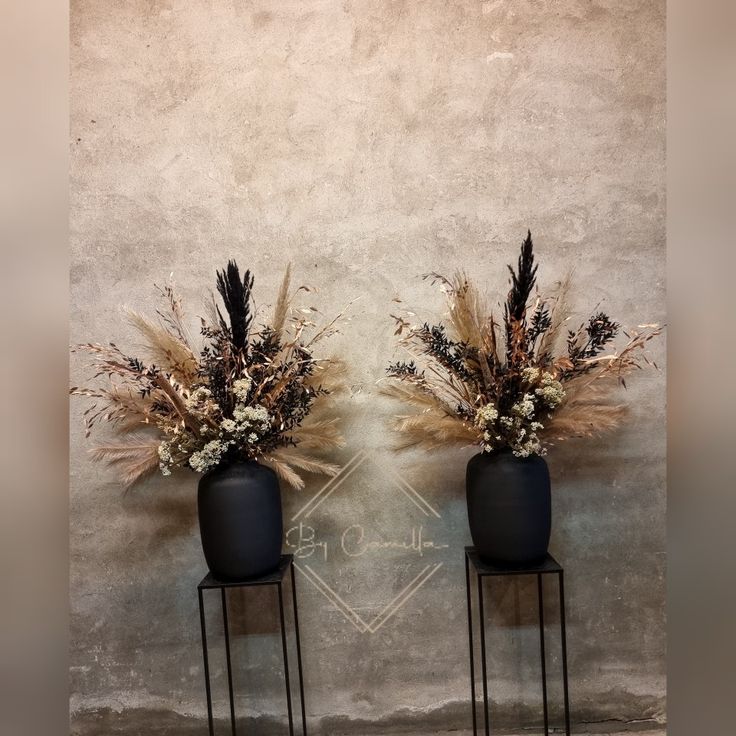 two black vases with dried flowers are on stands against a concrete wall, one is empty and the other has dry grass in it