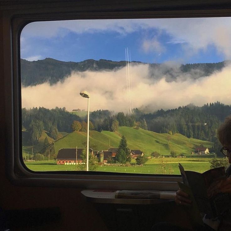 a person sitting on a train reading a book in front of a window with mountains and clouds behind them