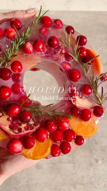 a person holding a doughnut with cranberries and oranges on it, surrounded by rosemary sprigs