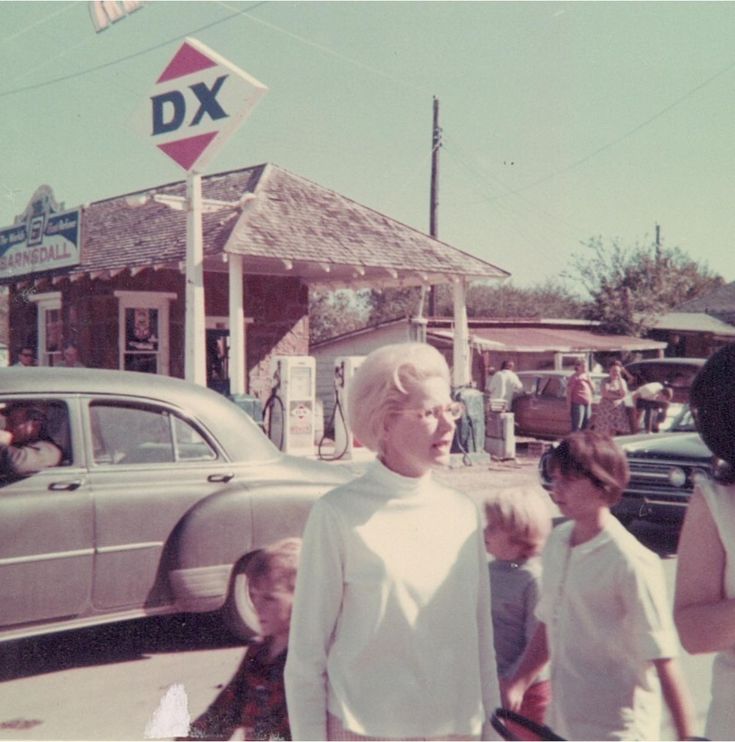 an older man standing in front of a car on the side of a road with people walking by