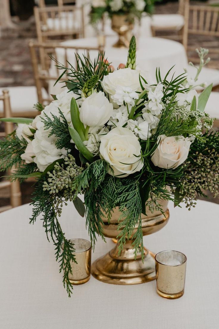 white flowers and greenery in a brass vase on a table with gold candlesticks