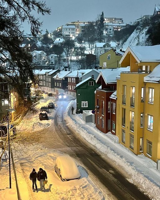 two people walking down a snow covered street in the middle of town at night time