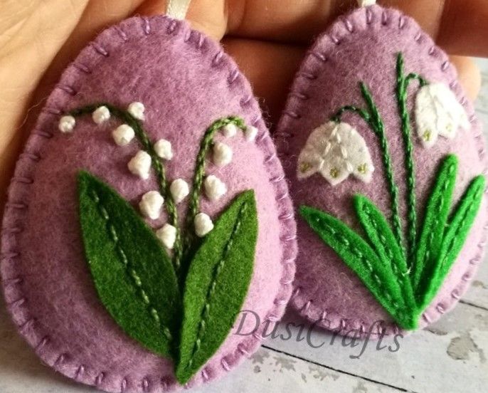 two handmade felt ornaments with white flowers and green leaves on purple background, one being held by a person's hand