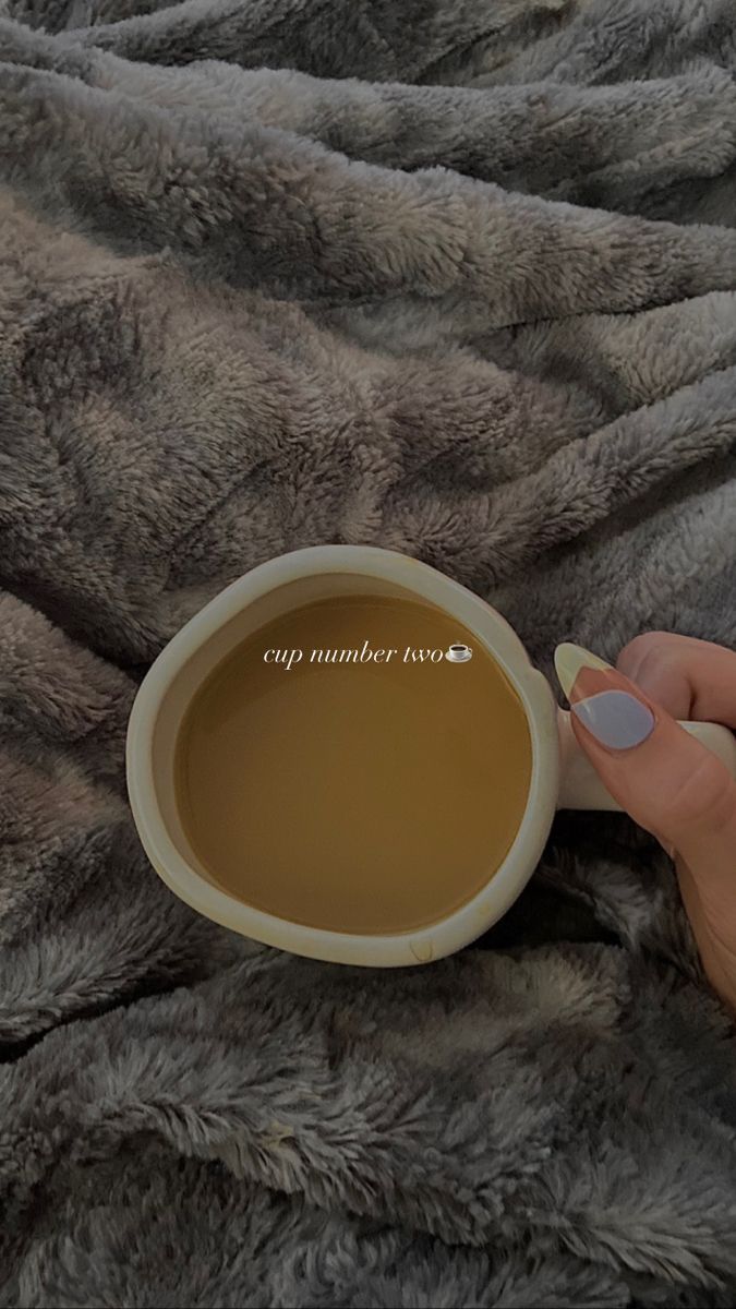 a woman holding a cup of coffee on top of a bed covered in grey blankets